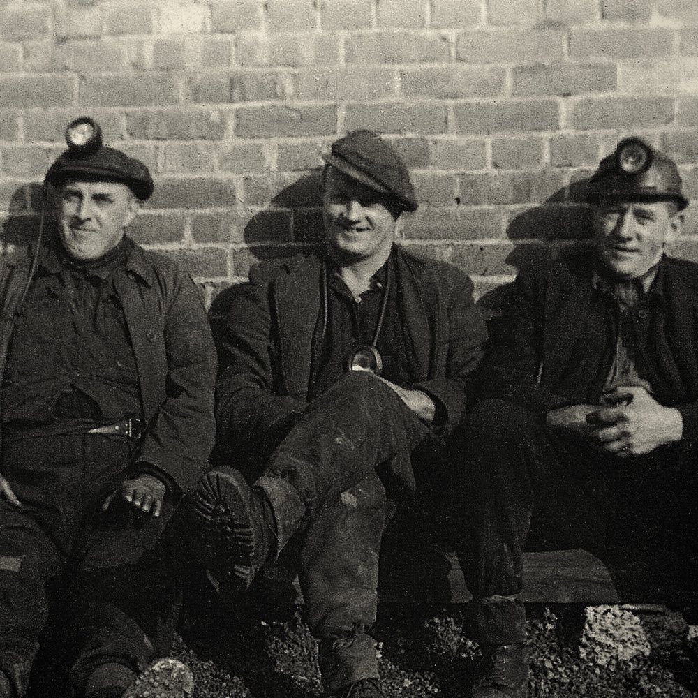 Three miners in caps and lamps, sitting against a brick wall in an old photograph