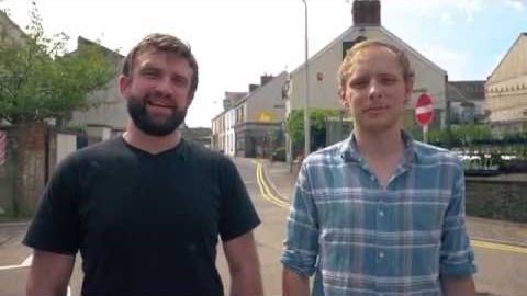 Two men standing in the middle of a Carmarthen street on a summer day, with terraced houses and a road sign behind them