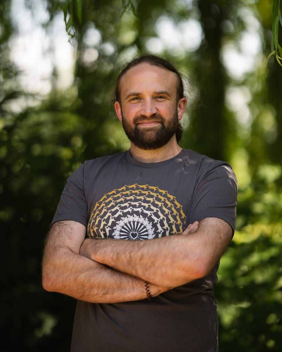 Deri outdoors among trees, arms folded, in a grey t-shirt