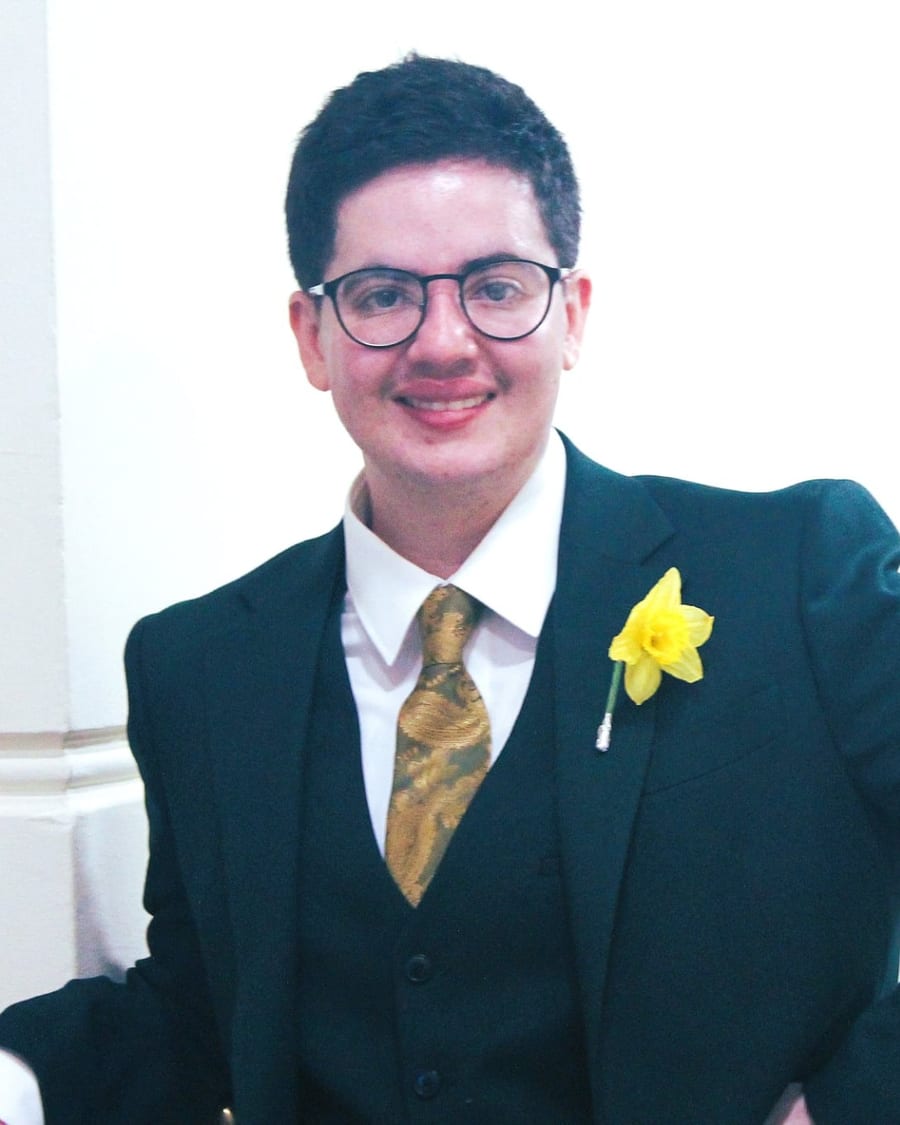Emrys in a dark three-piece suit with a gold tie and a daffodil, smiling