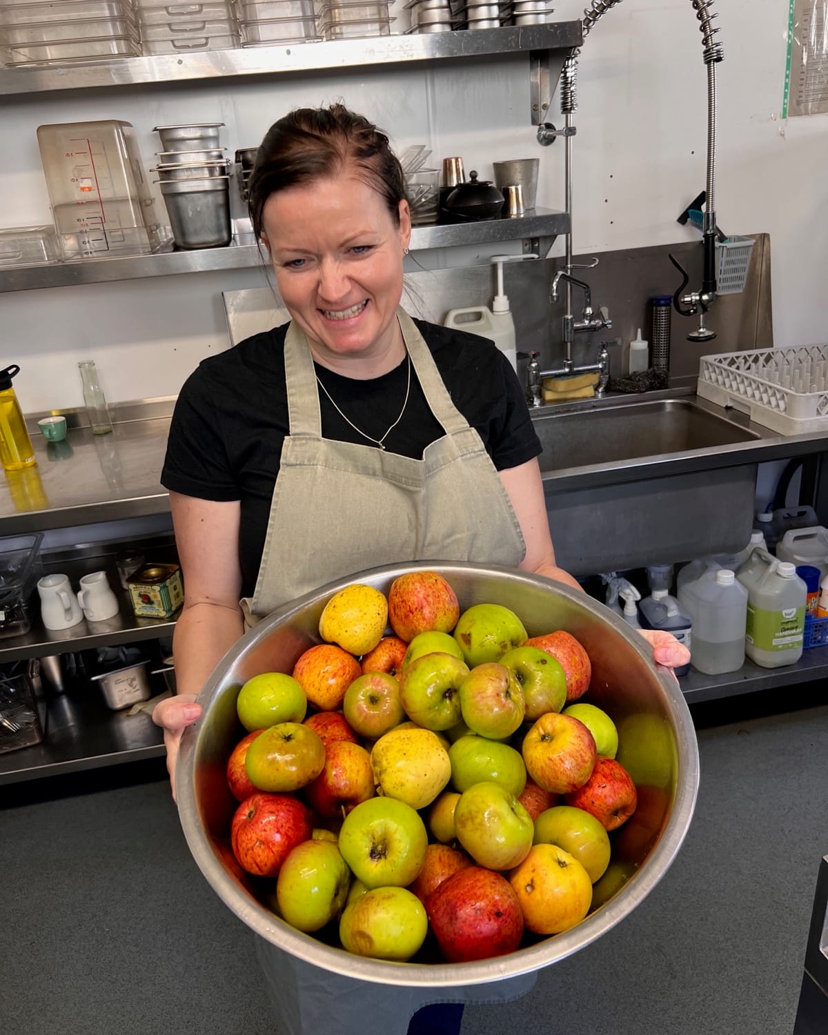 Tracy in an apron in the kitchen, laughing, holding a big bowl of apples